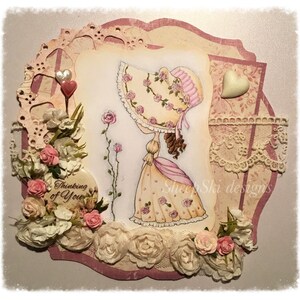 May include: A handmade card with a pink and white floral design. The card features a girl in a pink dress and a white hat, with the words "Thinking of You" and "Sheep Ski Designs" on the card.