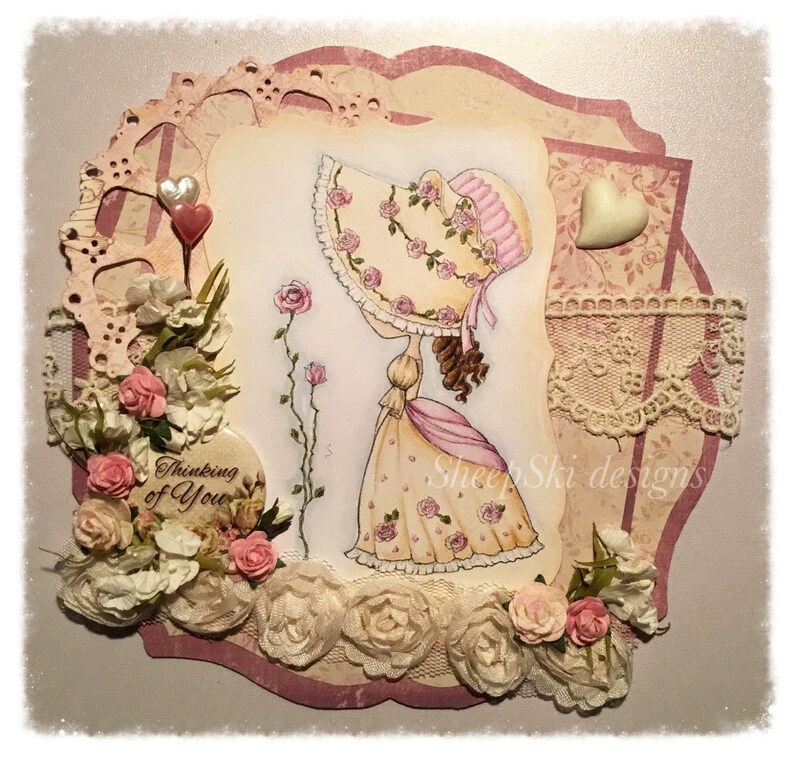 May include: A handmade card with a pink and white floral design. The card features a girl in a pink dress and a white hat, with the words "Thinking of You" and "Sheep Ski Designs" on the card.