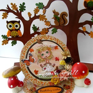 May include: A handmade autumn-themed birthday card with a layered design. The card features a whimsical girl with a butterfly, a squirrel, owls, and mushrooms. The card is decorated with fall leaves, flowers, and a brown banner with the text "Birthday wishes".