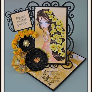 May include: A handmade card with a black frame and a yellow background. The card features a cartoon girl with long brown hair and a yellow flower crown. The card also includes a black and yellow flower embellishment and a sign that says "Hugs, Kisses and lots of Good Wishes!"
