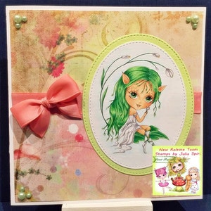 May include: A greeting card with a pink bow and a green fairy with green hair holding a white flower. The card has a green oval with a white background and a green border. The card has a pink and green background with white dots. The card has a small square with a picture of three fairies and the text "New Release Team Stamps by Julia Spiri Ann-Marie".