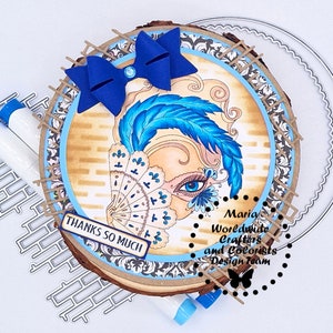 May include: A wood slice with a blue and white masquerade mask design, a blue bow, and the text "Thanks so much". The wood slice is surrounded by a decorative border and the text "Maria Worldwide Crafters and Colorists Design Team".