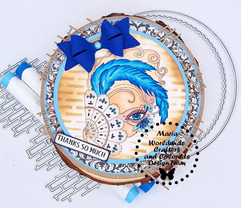 May include: A wood slice with a blue and white masquerade mask design, a blue bow, and the text "Thanks so much". The wood slice is surrounded by a decorative border and the text "Maria Worldwide Crafters and Colorists Design Team".