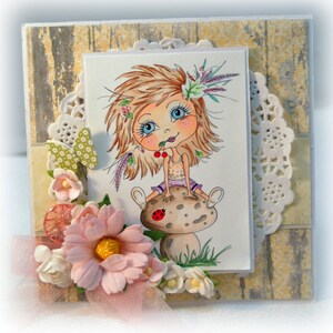 May include: A handmade card featuring a cartoon girl with blonde hair sitting on a mushroom. The girl is wearing a purple dress and holding a red cherry. The card is decorated with flowers, lace, and a wooden background.