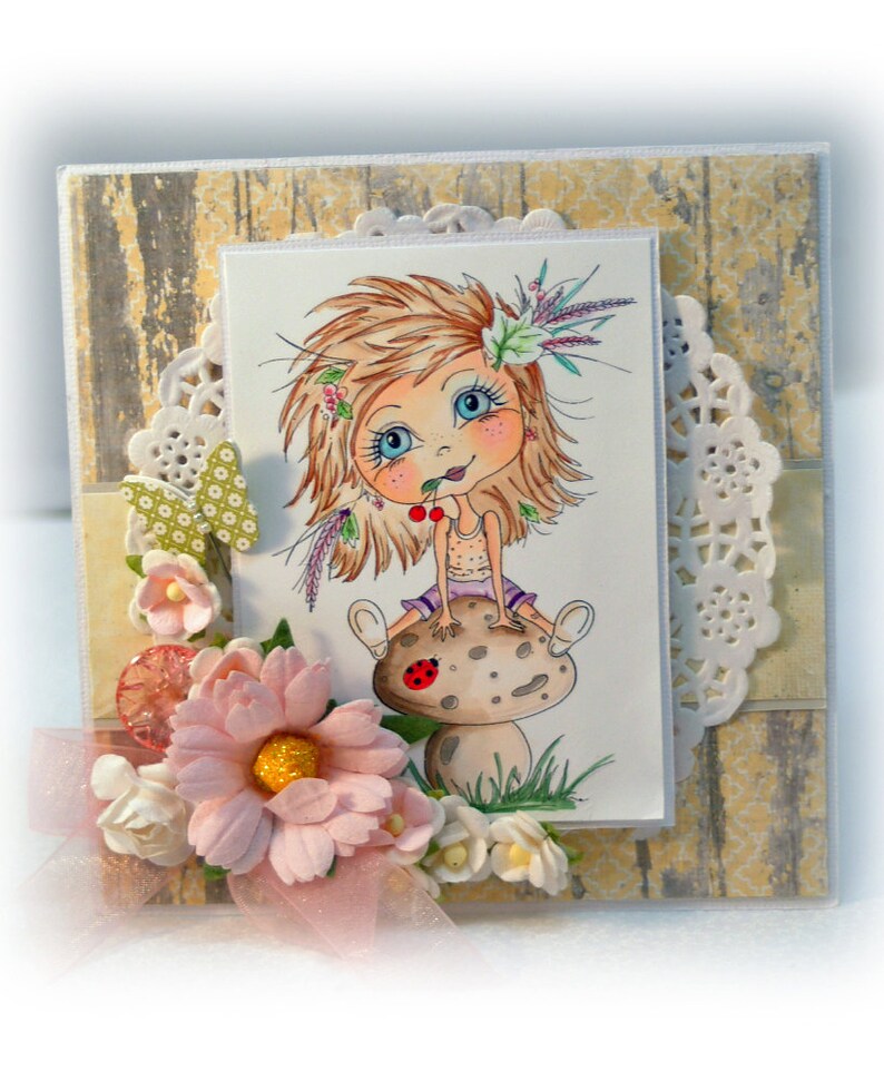 May include: A handmade card featuring a cartoon girl with blonde hair sitting on a mushroom. The girl is wearing a purple dress and holding a red cherry. The card is decorated with flowers, lace, and a wooden background.