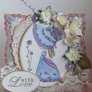 May include: A handmade card with a pink background and a white oval in the center. The oval features a drawing of a girl in a blue dress and a hat, with flowers and vines around her. The card also has a white oval at the bottom with the words "with love" written in cursive.