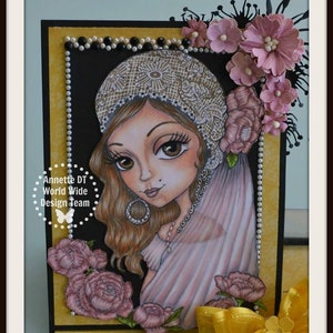 May include: A card featuring a hand-drawn illustration of a woman with long brown hair wearing a white lace hat and a pink dress. The woman is surrounded by pink flowers and a yellow bow. The card has a black background with a gold border and the text "Annette DT World Wide Design Team" in a circle.