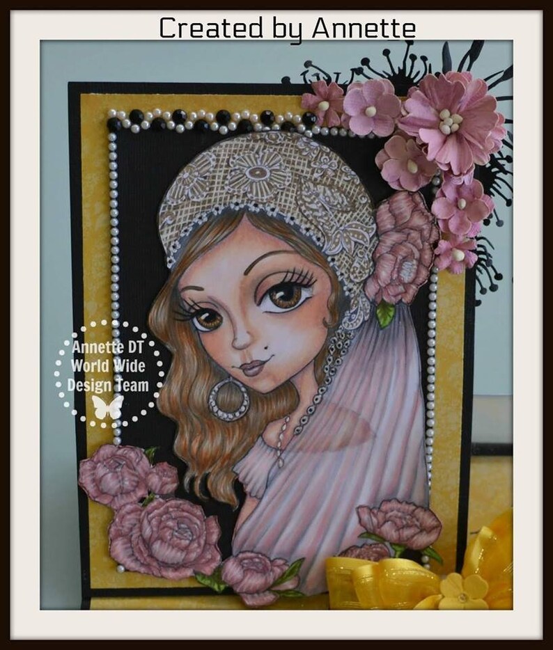 May include: A card featuring a hand-drawn illustration of a woman with long brown hair wearing a white lace hat and a pink dress. The woman is surrounded by pink flowers and a yellow bow. The card has a black background with a gold border and the text "Annette DT World Wide Design Team" in a circle.