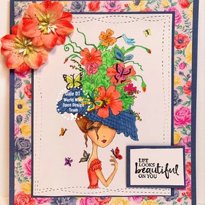 May include: A colorful greeting card with a whimsical illustration of a woman wearing a hat adorned with flowers and butterflies. The text "Life looks beautiful on you" is printed on the card.