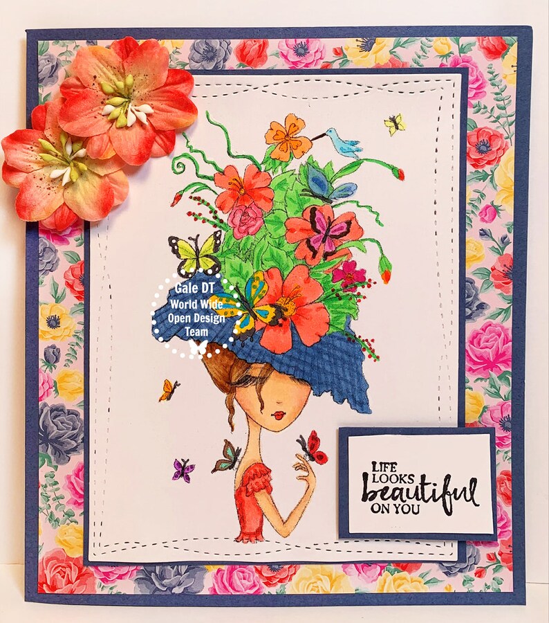 May include: A colorful greeting card with a whimsical illustration of a woman wearing a hat adorned with flowers and butterflies. The text "Life looks beautiful on you" is printed on the card.