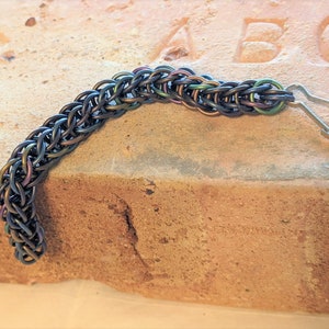 May include: A handcrafted chainmail bracelet with a lobster clasp. The bracelet is made of small, interlocking rings in shades of iridescent blue, green, and purple. The chainmail is curved and rests on a textured, light brown surface.
