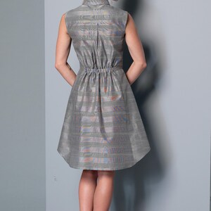 May include: Sleeveless gray dress with a collared neckline and elastic waist. The dress features a subtle striped pattern with hints of orange and blue. The skirt flares out from the waist. The model is wearing black flats.