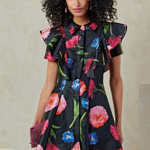Puede incluir: Un vestido negro de estampado floral con cuello de volantes y mangas cortas. El vestido tiene un cierre de botones en la parte delantera y una falda acampanada.