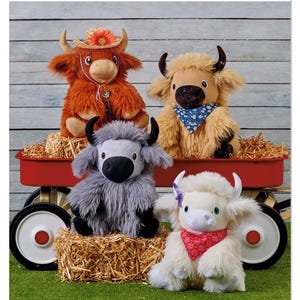 May include: Simplicity pattern S3221 features four plush Highland cow toys in a red wagon and on hay bales. The cows are in shades of brown, gray, and white, with accessories like bandanas and a hat. The image includes the Simplicity logo and "Designed and printed in USA."