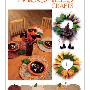 May include: A set of five pumpkin-shaped placemats with different patterns, including red, orange, brown, and white. The placemats are arranged in a row and are labeled with the number M7490. The image also shows a witch-themed wreath and a table setting with witch-themed decorations.