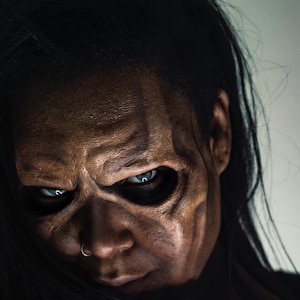 May include: A close-up of a person's face with dramatic makeup, featuring dark brown and tan tones to create a skeletal effect. The eyes are piercing blue, and the person has a nose ring. Long black hair frames the face.