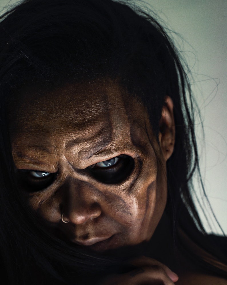 May include: A close-up of a person's face with dramatic makeup, featuring dark brown and tan tones to create a skeletal effect. The eyes are piercing blue, and the person has a nose ring. Long black hair frames the face.