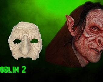 May include: A close-up of a goblin mask, showing the detailed features of the mask, including the large nose, pointed ears, and wrinkled skin. The mask is made of a light-colored material and is ready to be painted. The text "GOBLIN 2" is visible below the mask.