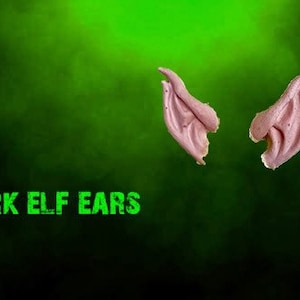 May include: A pair of dark elf ears, made of a soft, flexible material. The ears are a light pink color and have a realistic, pointed shape. The text "DARK ELF EARS" is printed in green below the ears.