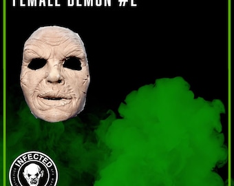 Peut inclure: Un gros plan d'un masque de démon féminin réaliste et détaillé avec une peau ridée et une expression sinistre. Le masque est placé sur un fond de fumée verte. Le texte "FEMALE DEMON #2" est visible en haut de l'image.