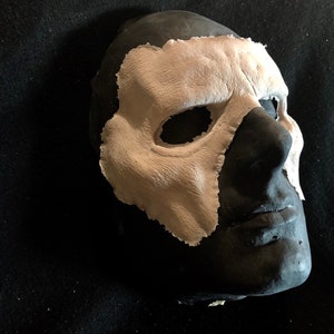 May include: A black mask with a textured, light beige overlay covering the right side of the face. The mask has eye and nose openings. The mask is likely for costume or theatrical use.