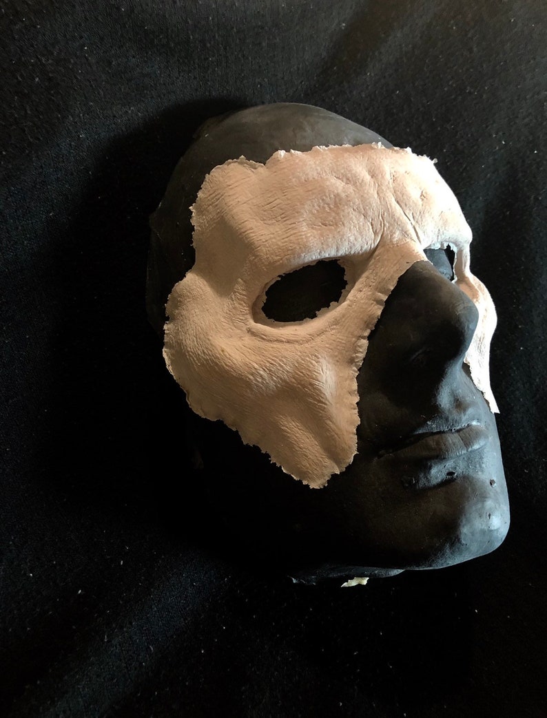 May include: A black mask with a textured, light beige overlay covering the right side of the face. The mask has eye and nose openings. The mask is likely for costume or theatrical use.