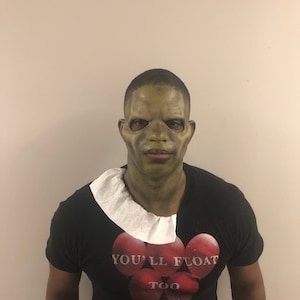 May include: A person wearing a green mask and a black t-shirt with the text "YOU'LL FLOAT TOO" printed on it. The mask has a grotesque appearance, and the shirt features a graphic of red balloons.