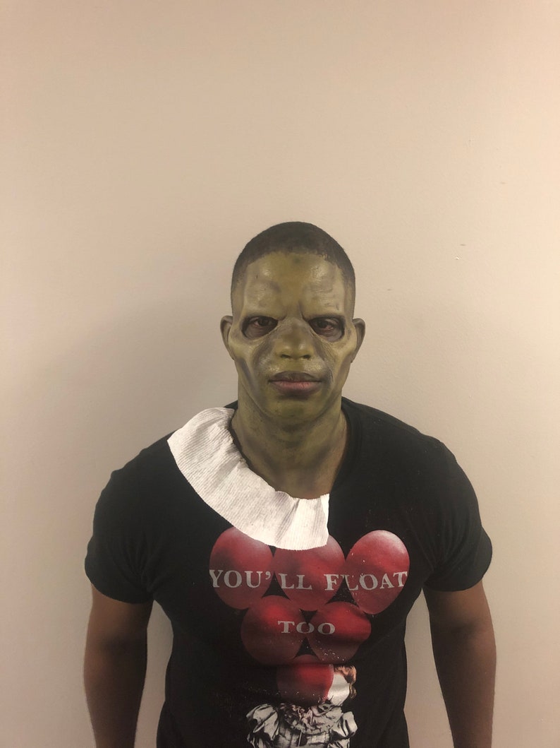 May include: A person wearing a green mask and a black t-shirt with the text "YOU'LL FLOAT TOO" printed on it. The mask has a grotesque appearance, and the shirt features a graphic of red balloons.