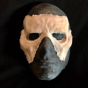 May include: A dark gray mask with a light tan eye covering. The mask has a textured appearance and is set against a black background. The eye covering has a rough, uneven edge.