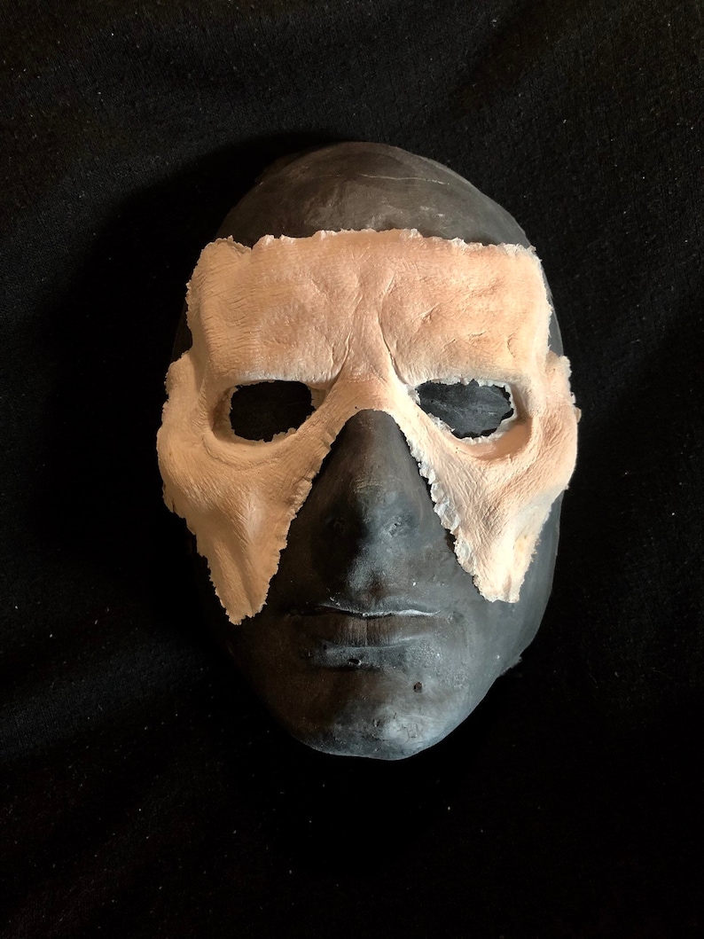 May include: A dark gray mask with a light tan eye covering. The mask has a textured appearance and is set against a black background. The eye covering has a rough, uneven edge.