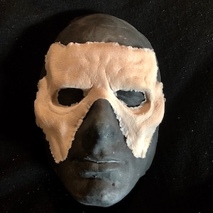 May include: A dark gray mask with a light beige eye covering. The mask has a textured surface and is set against a black background. The eye covering has a rough, uneven edge.