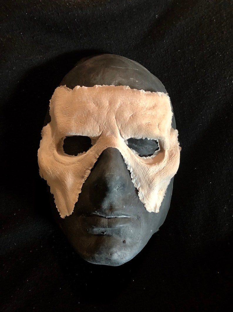 May include: A dark gray mask with a light beige eye covering. The mask has a textured surface and is set against a black background. The eye covering has a rough, uneven edge.