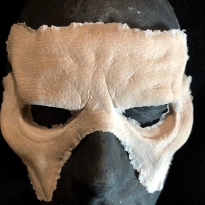 May include: A light beige mask with eye and nose openings, set against a dark background. The mask has a textured surface and a rough, torn edge, suggesting a handmade or aged appearance. The mask is likely for costume or theatrical use.