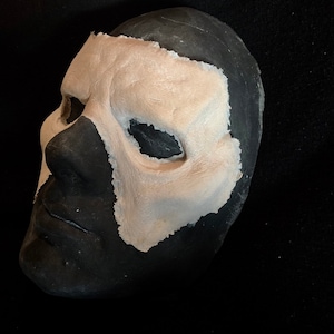 May include: A black and beige mask with a textured surface. The mask is painted black on the lower half and beige on the upper half, with eye and nose openings. The mask is likely for costume or theatrical use.