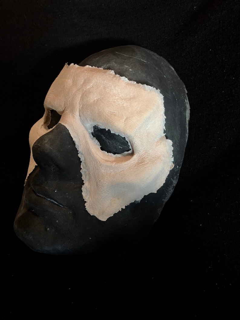 May include: A black and beige mask with a textured surface. The mask is painted black on the lower half and beige on the upper half, with eye and nose openings. The mask is likely for costume or theatrical use.
