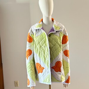 May include: A quilted jacket with a unique design. The jacket features a light green and white pattern with orange leaf-like shapes. It has a purple lining and an orange collar. The jacket is displayed on a mannequin.