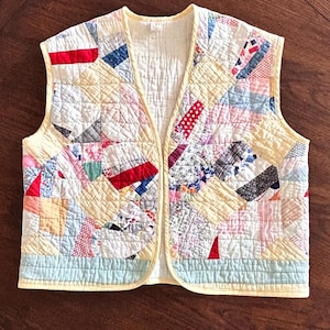 May include: A handmade, quilted vest with a patchwork design. The vest features a variety of fabric patterns in red, blue, pink, and white, set against a pale yellow background. The vest has a light blue border and a yellow trim.