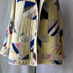 May include: A hooded jacket constructed from a patchwork quilt featuring a variety of fabric patterns in shades of yellow, blue, pink, and white. The garment includes two large square front pockets and is finished with cream-colored trim.
