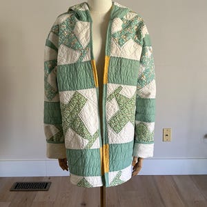 May include: A quilted jacket with a hood, displayed on a mannequin. The jacket features a patchwork design in shades of green, white, and yellow. The jacket is open in the front, revealing the yellow lining. The overall style is vintage and handmade.