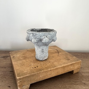May include: A handmade, textured, light gray and white vase with a wide top and a narrow base. The vase has a rough, organic appearance and sits on a wooden, rectangular stand. The background is a plain white wall.
