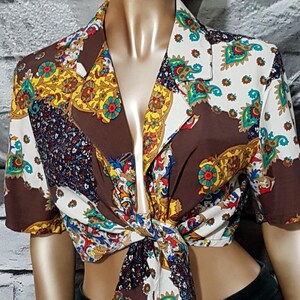 Op de afbeelding: Een blouse met korte mouwen en een patroon met een strik aan de voorkant. De blouse heeft een mix van kleuren, waaronder bruin, wit, geel en blauw. De stof heeft een vintage uitstraling, met een patchwork-achtig patroon. De blouse wordt op een mannequin getoond.