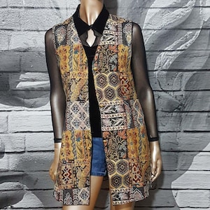 Op de afbeelding: Een lange, mouwloze vest met een patchwork-ontwerp in tinten bruin, geel en blauw. De vest heeft een knoopsluiting en een kraag.