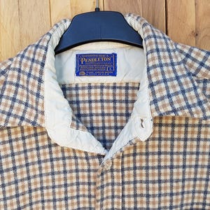 Peut inclure: Une chemise à manches longues boutonnée avec un motif à carreaux beige et bleu foncé. La chemise présente une doublure de col intérieure de couleur crème et une étiquette de marque Pendleton bleue cousue au niveau du cou sur un cintre noir.