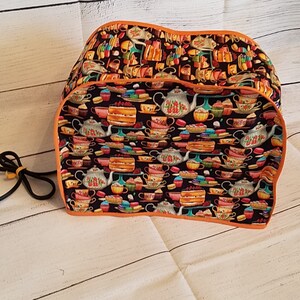 May include: A black and orange toaster cover with a colorful pattern of teacups, teapots, and cakes. The cover has an orange trim and a black cord with a yellow clasp.