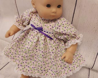 Puede incluir: Una muñeca con un vestido floral rosa con detalles morados y ribete de encaje blanco. El vestido tiene mangas cortas y una cinta morada. La muñeca tiene tez clara y está sobre una superficie de madera blanca.