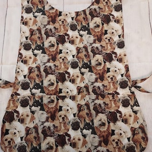 May include: An apron featuring a repeating pattern of various dog breeds in shades of brown, tan, and white. The apron has a bib top, a large pocket, and ties at the waist. The background is a light wood grain.