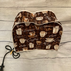 May include: A brown and white coffee maker cover with a repeating pattern of coffee cups, coffee beans, and coffee-related words. The cover has a white trim and a black power cord.