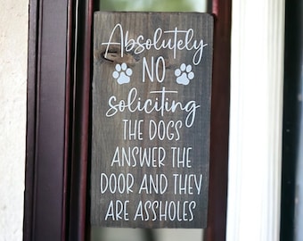 Könnte beinhalten: Rechteckiges Holzschild mit weißem Text: "Absolutely NO soliciting THE DOGS ANSWER THE DOOR AND THEY ARE ASSHOLES". Zwei Pfotenabdrücke über dem Wort "NO". Das Holz ist dunkel gebeizt.