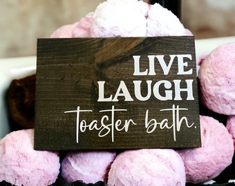 Może przedstawiać: Drewniany znak z napisem "Live Laugh Toaster Bath" w białych literach na ciemnobrązowym tle. Znak jest otoczony różowymi bombami kąpielowymi.
