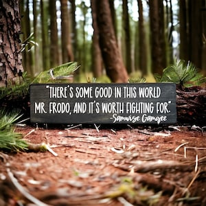 May include: A black wooden sign with white text that reads "THERE'S SOME GOOD IN THIS WORLD MR. FRODO, AND IT'S WORTH FIGHTING FOR." -Samwise Gamgee. The sign is lying on the ground in a forest.